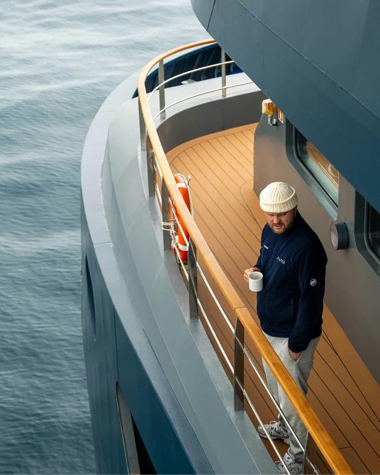 Crew member in a knitted beanie on the curved teak side deck of a superyacht, holding a mug, grey water below.